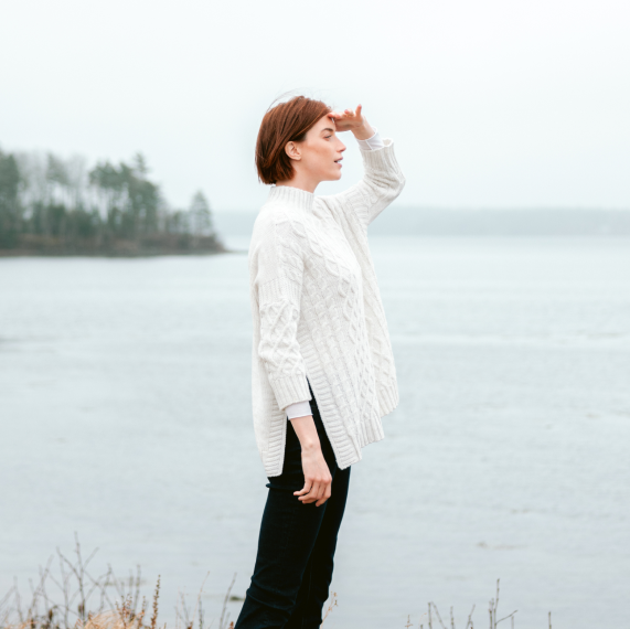 female wearing white sweater and dark pants looking out to the water off the coast