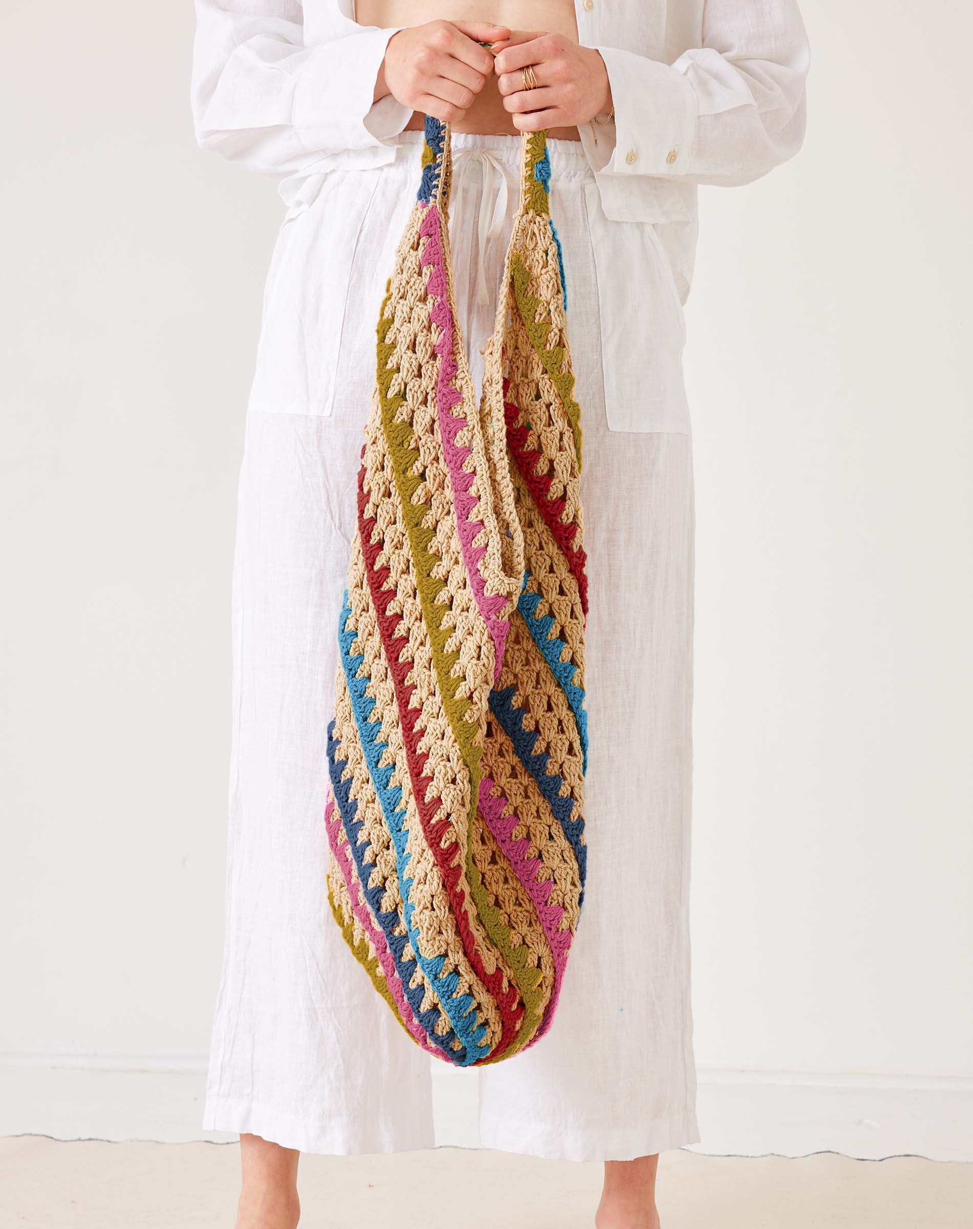 Colorful crocheted bag held by a person wearing a white outfit on a plain background