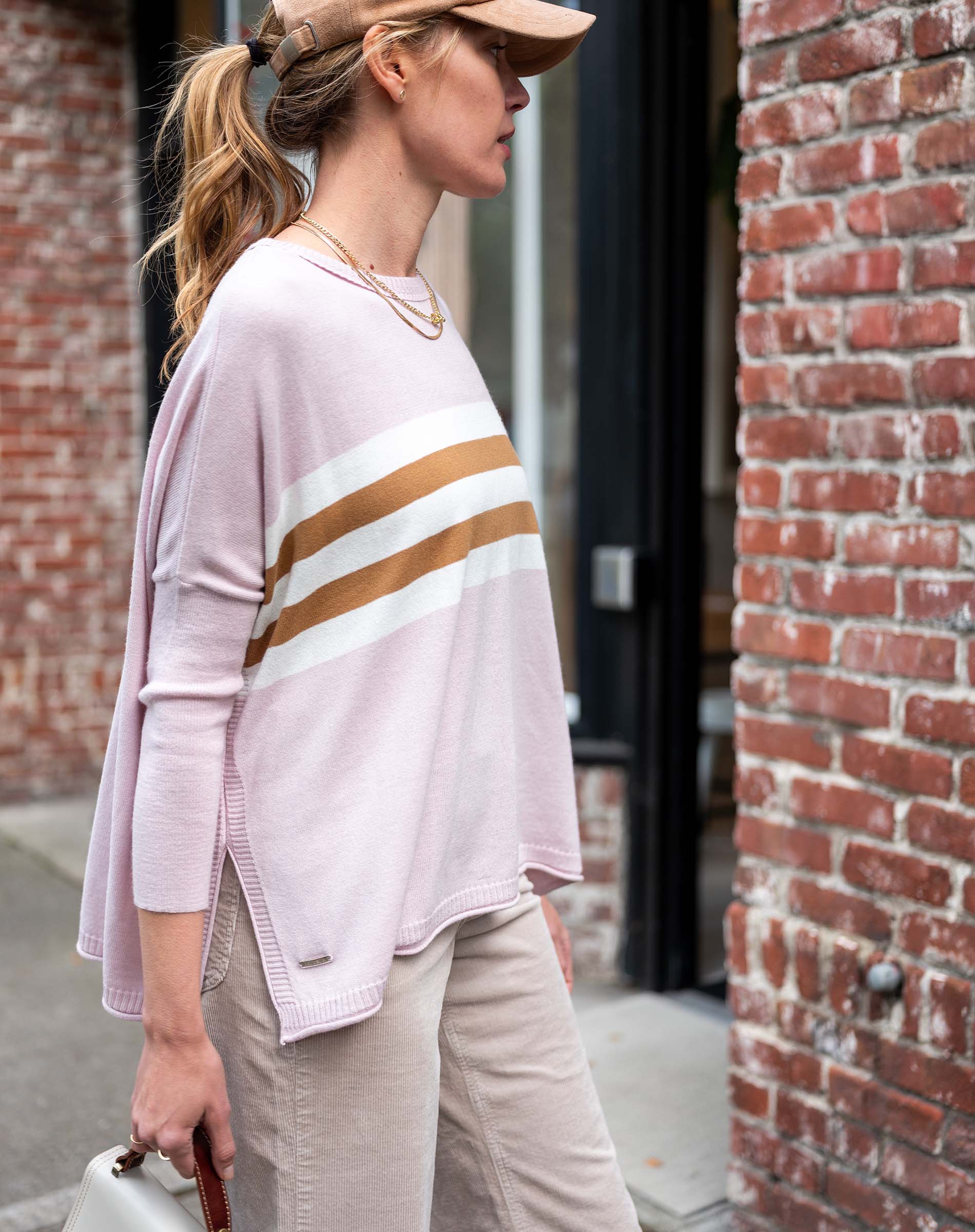 Woman wearing a light pink sweater with gold stripes and beige pants, standing against a brick wall.