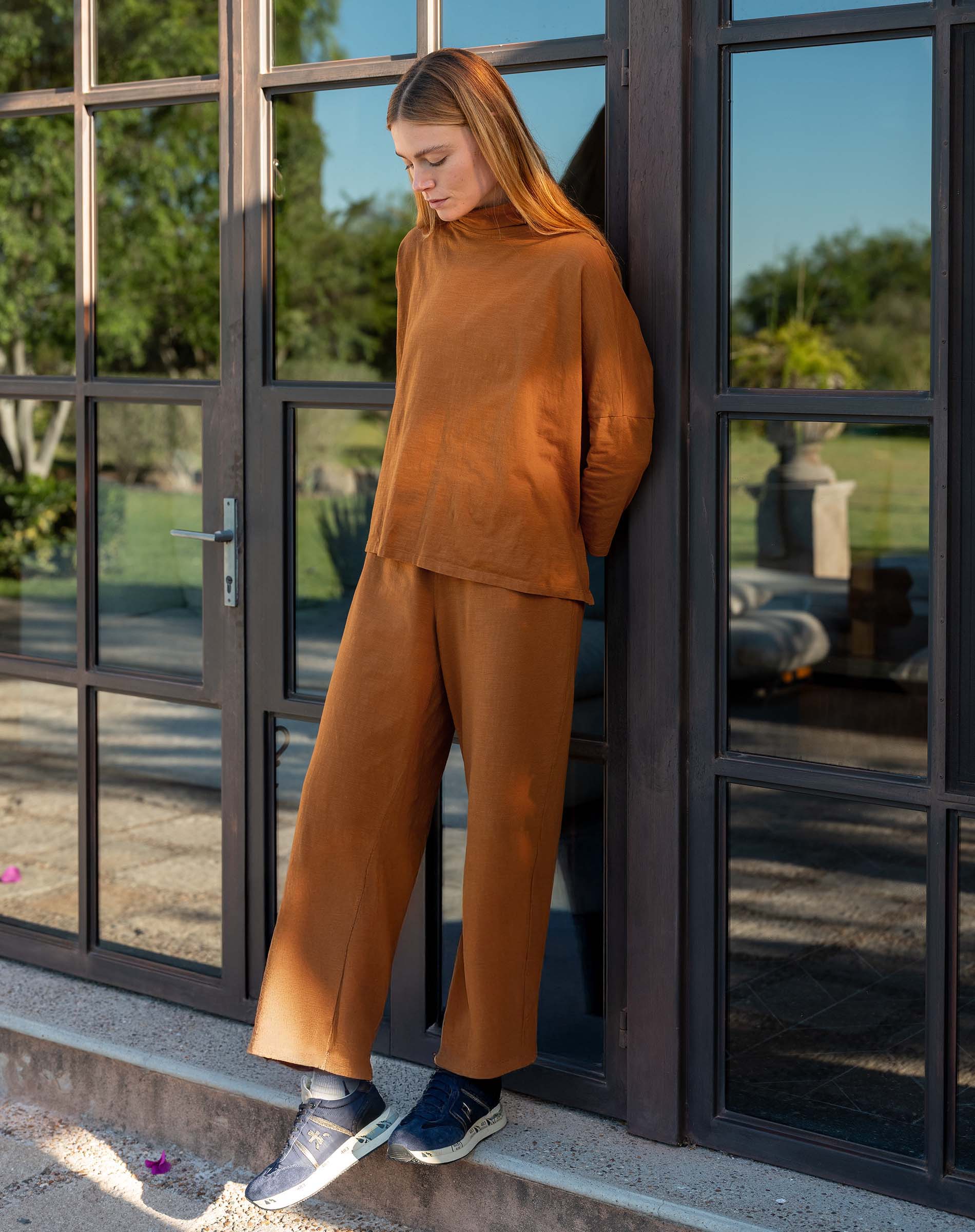Person wearing a brown outfit standing by a glass door with a garden view.
