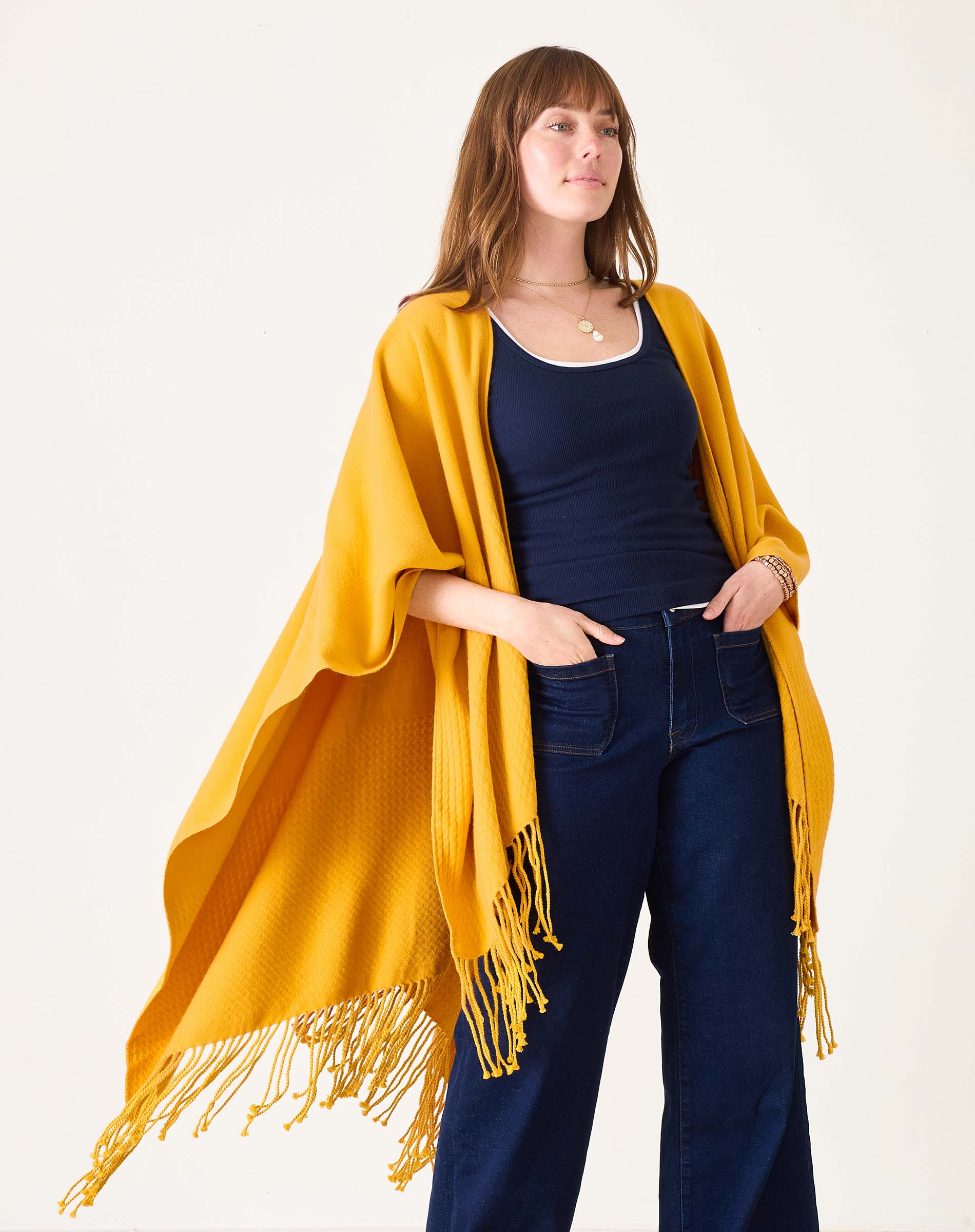 Woman wearing a yellow fringed poncho over a dark top and jeans on a white background