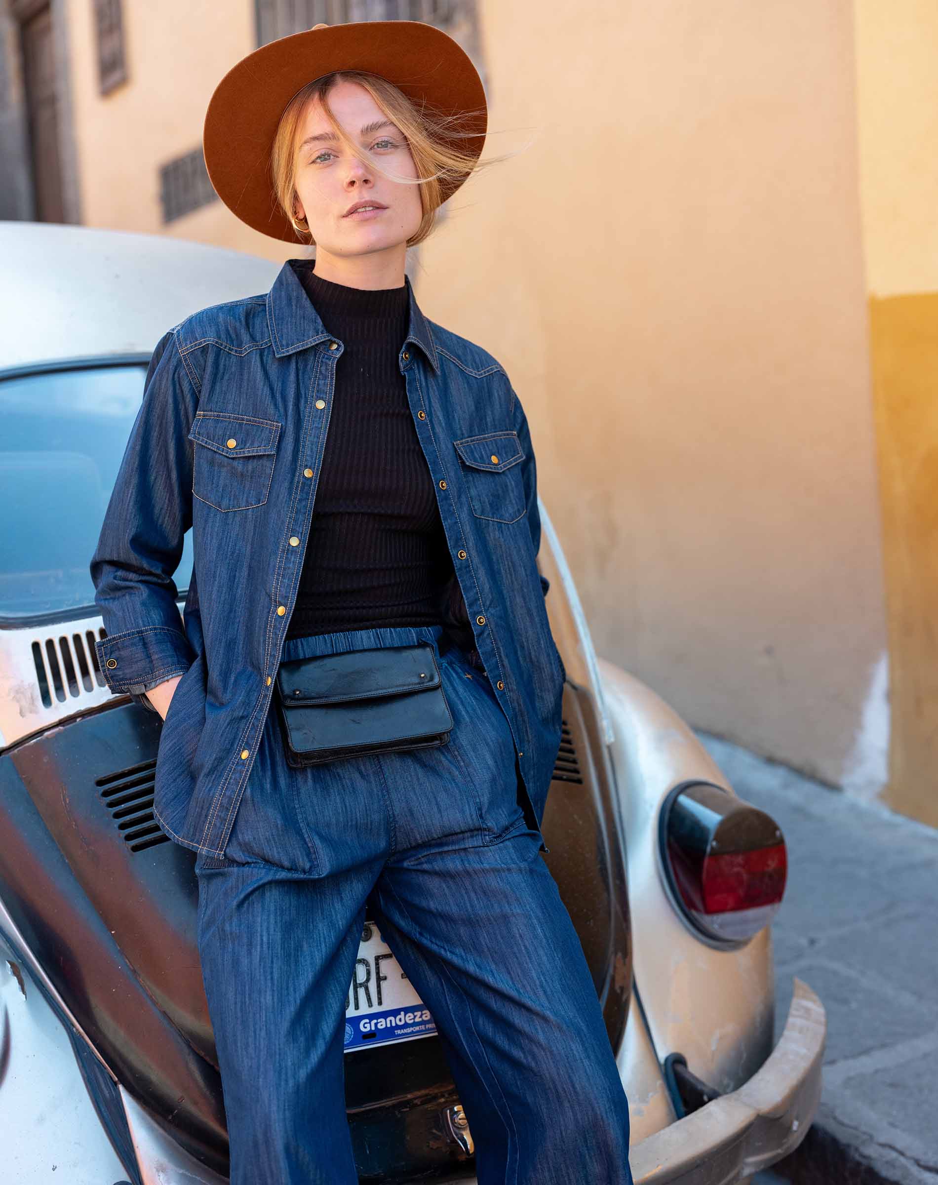 Person wearing a blue denim outfit and brown hat leaning against a vintage car.