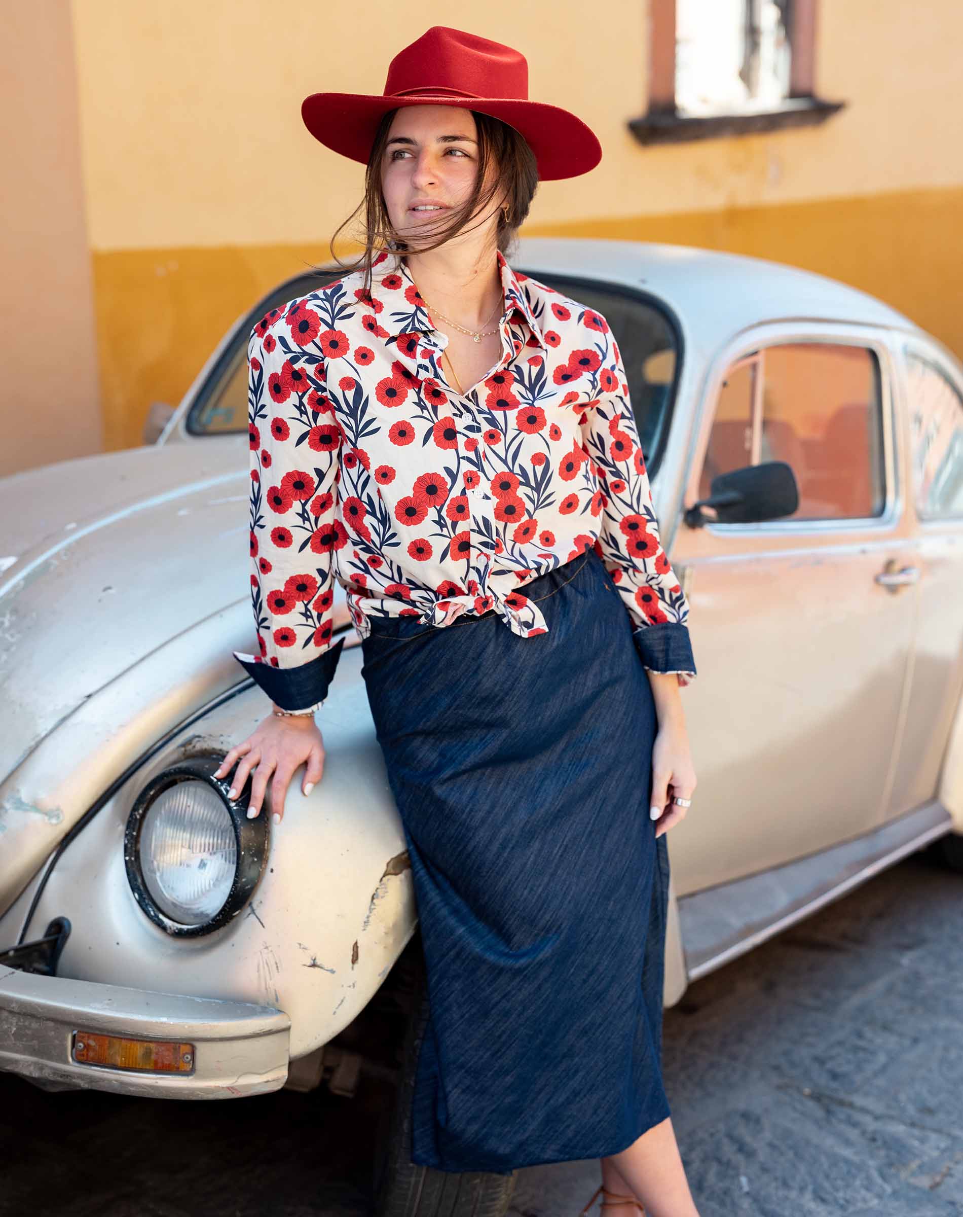 Woman in a floral blouse and red hat standing next to a vintage car.