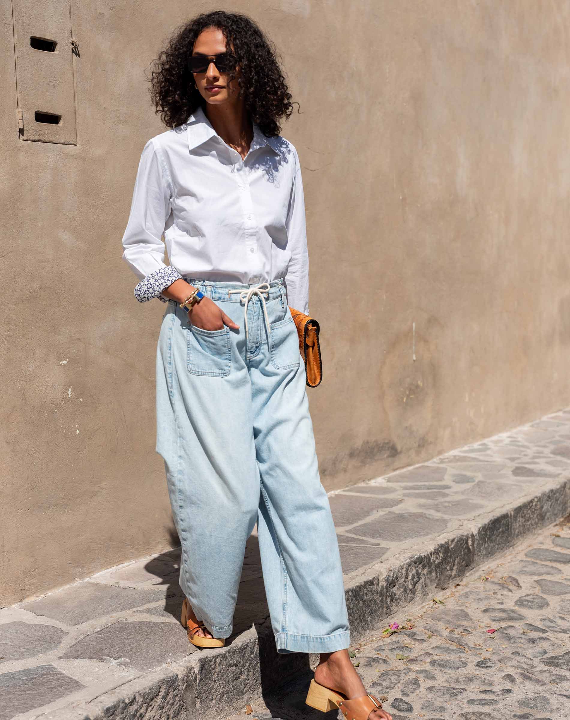 Woman wearing a white shirt and light blue jeans standing on a stone sidewalk.