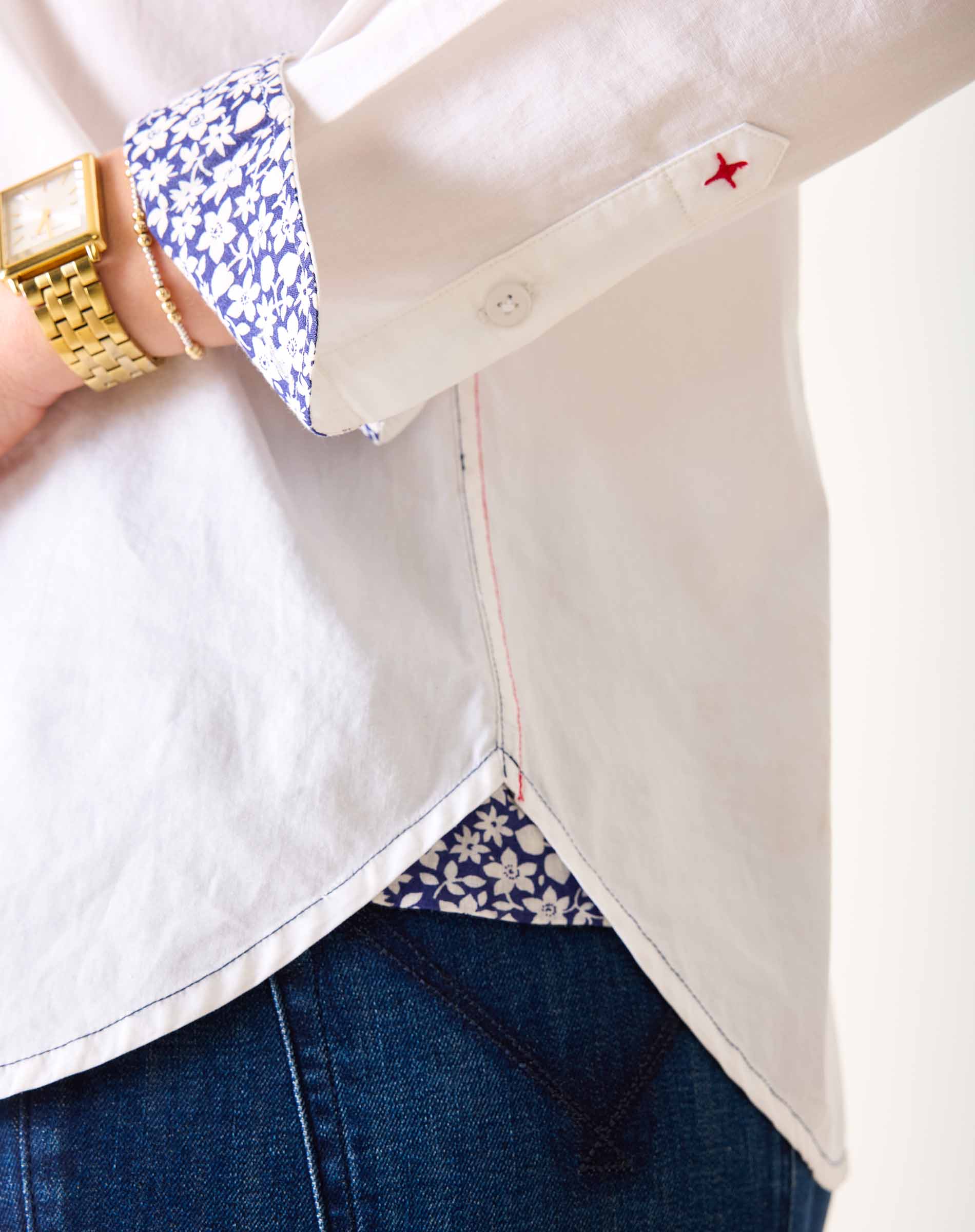 Close-up of a person wearing a white shirt with floral cuffs, blue jeans, and a gold watch.