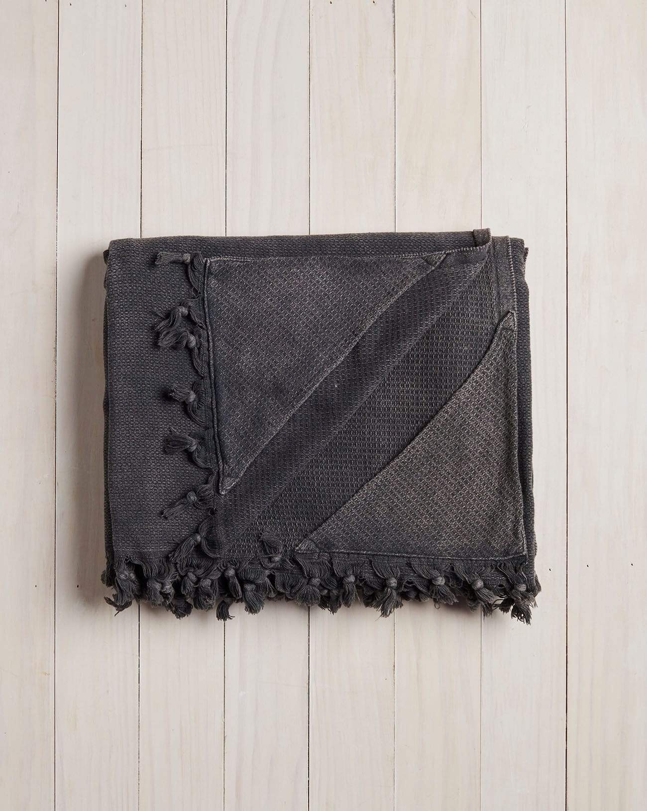 folded dark grey beach blanket with fringe on a white wood background