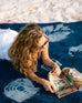 female laying on blue shibori blanket flipping through a magazine laying down on the beach