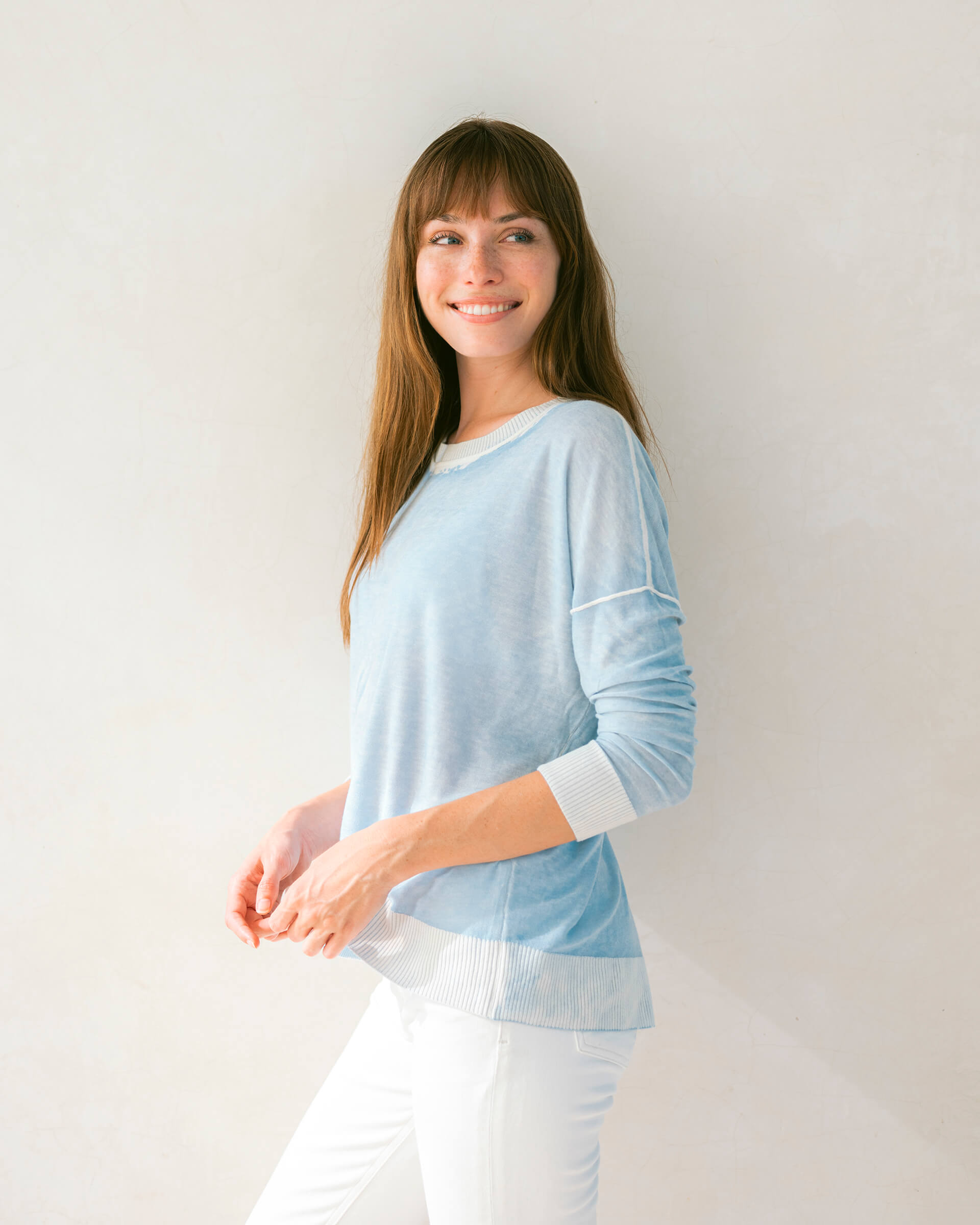 brunette female wearing light blue sweater standing sideways in front of a white wall 