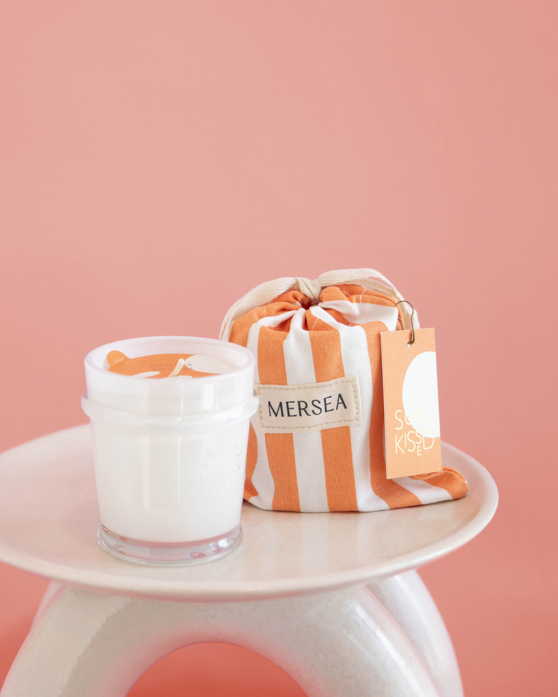 6.5 oz Sun Kissed sandbag candle in orange and white striped bag on a light red background