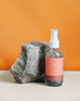 4 oz clear glass bottle of Sun Kissed room spray next to a rock on an orange background