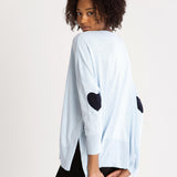 rearview of female wearing light blue sweater with blue  heart elbow patch on white background