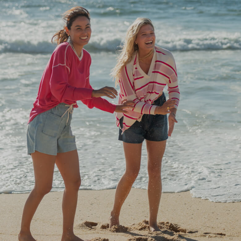 two women having fun on the beach wearing sweaters and jean shorts