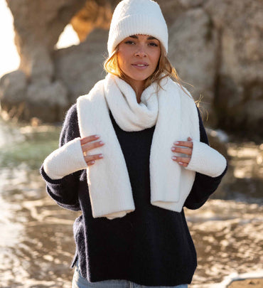 female wearing matching white beanie, fingerless gloves, and scarf over a navy sweater on the beach