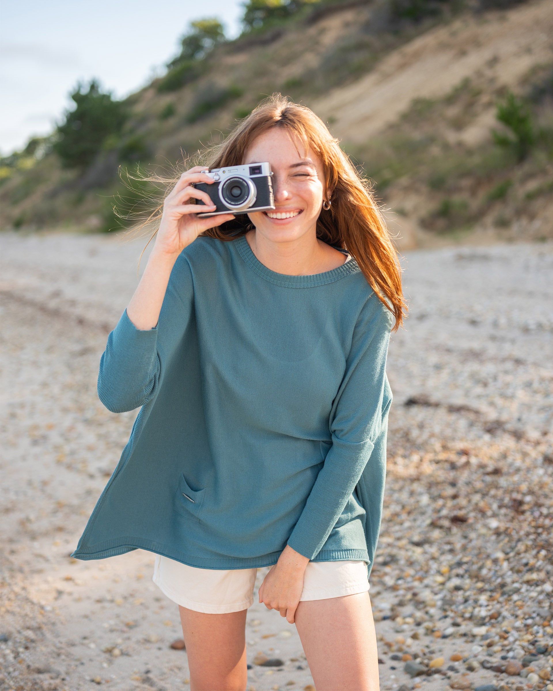 female wearing greenish blue sweater standing on the beach holding a camera
