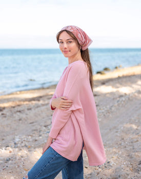 brunette female wearing pink sweater standing sideways on a beach with a hand to her elbow