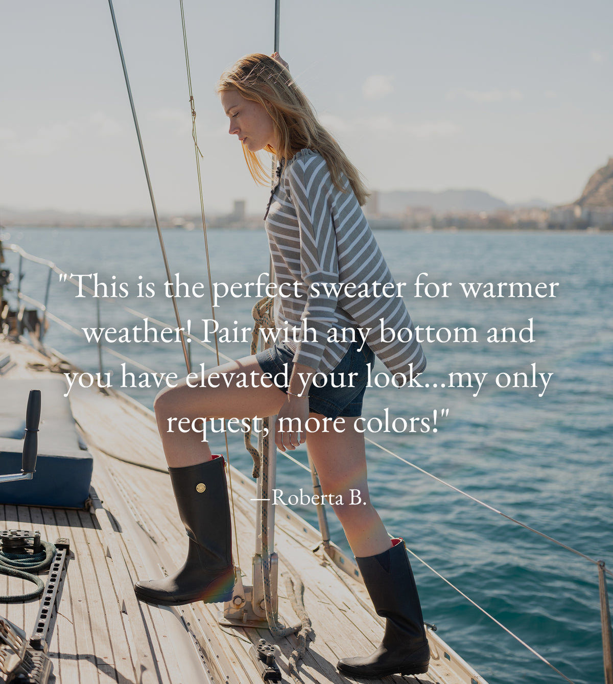 A woman standing on a wooden boat deck overlooking a coastal shoreline, wearing a striped sweater and boots. Text: "This is the perfect sweater for warmer weather! Pair with any bottom and you have elevated your look...my only request, more colors!" —Roberta B.