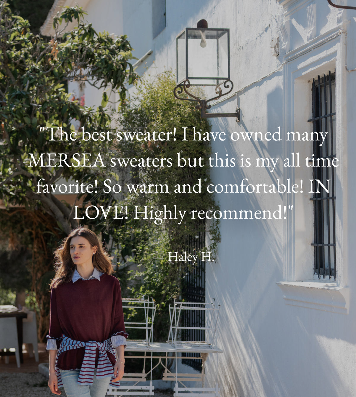 A woman standing in a Mediterranean-style courtyard wears a burgundy long-sleeved shirt with a striped scarf tied around her waist. Overlaying the image is a customer review: "The best sweater! I have owned many MERSEA sweaters but this is my all time favorite! So warm and comfortable! IN LOVE! Highly recommend!" - Haley H.