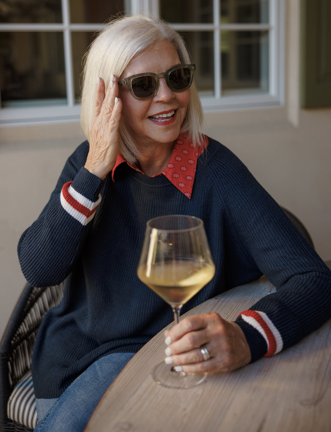 A woman with blonde hair and sunglasses smiles while holding a glass of white wine at an outdoor table.