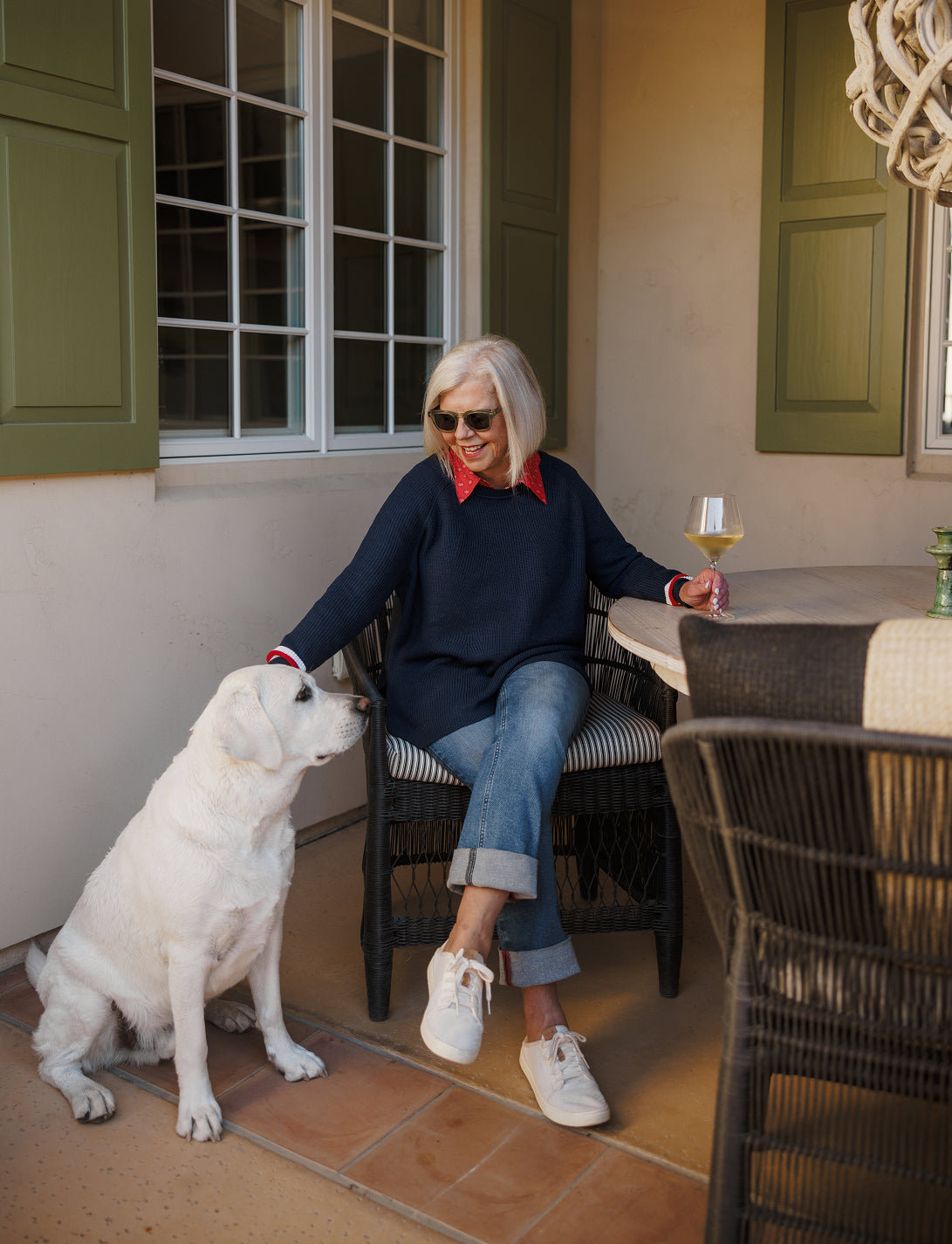 A woman with blonde hair and sunglasses sits on a patio chair next to a large white dog, holding a glass of wine. She is wearing a navy blue knit sweater over a red collared shirt and denim jeans.