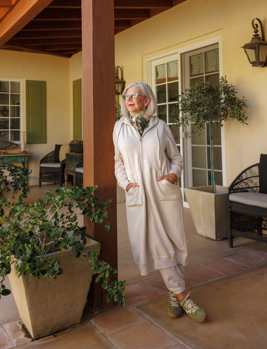 A woman wearing a long cream-colored tunic and sunglasses stands on a patio in front of large glass doors.
