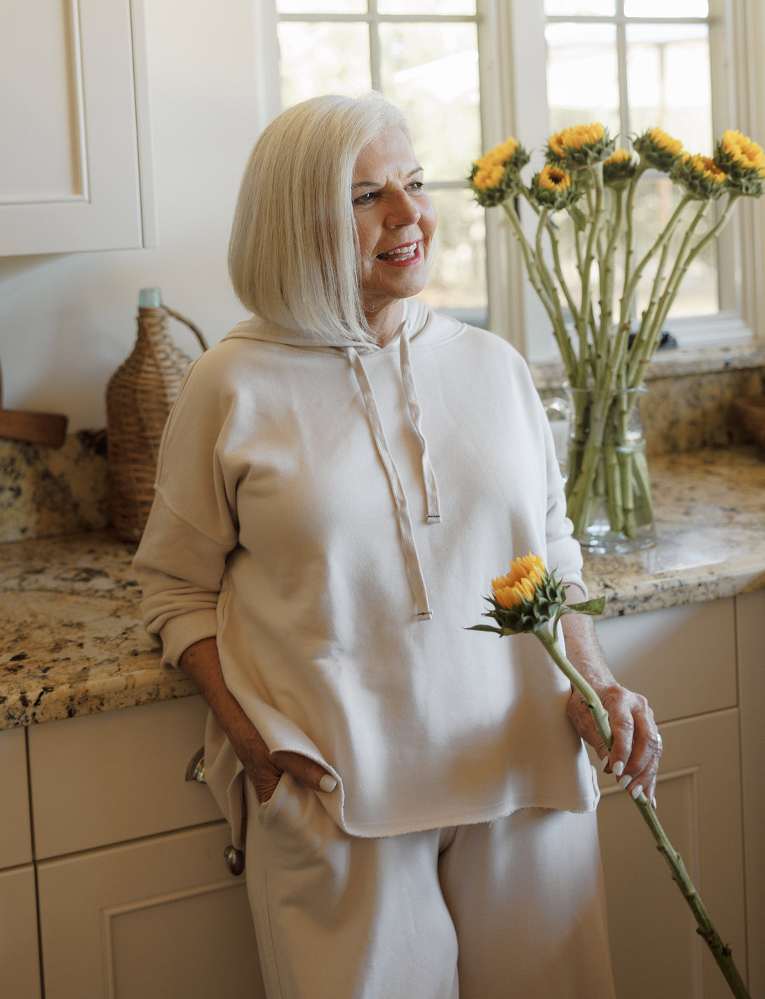 A woman with short blonde hair wearing a cream-colored knit hoodie and matching pants stands in a kitchen holding a single yellow flower near a vase of flowers.