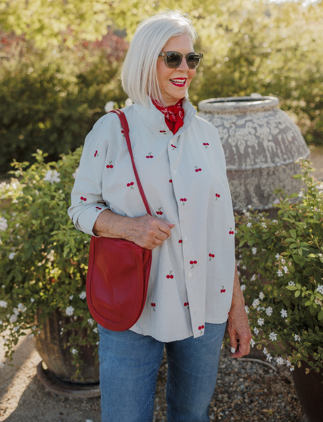 A woman with short white hair and sunglasses smiles while wearing a cherry print blouse and red scarf in a garden setting.