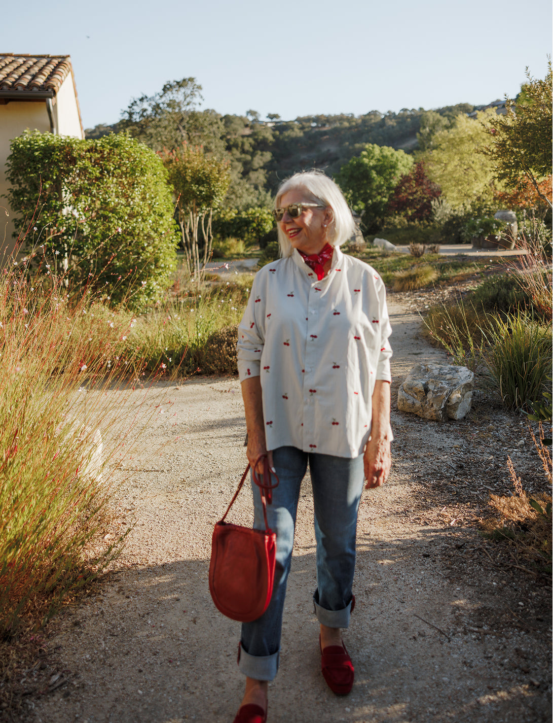 A woman with white hair and sunglasses stands outdoors in a sun-drenched garden wearing a button-down shirt with a small floral print, denim jeans, and red flats, holding a leather handbag.