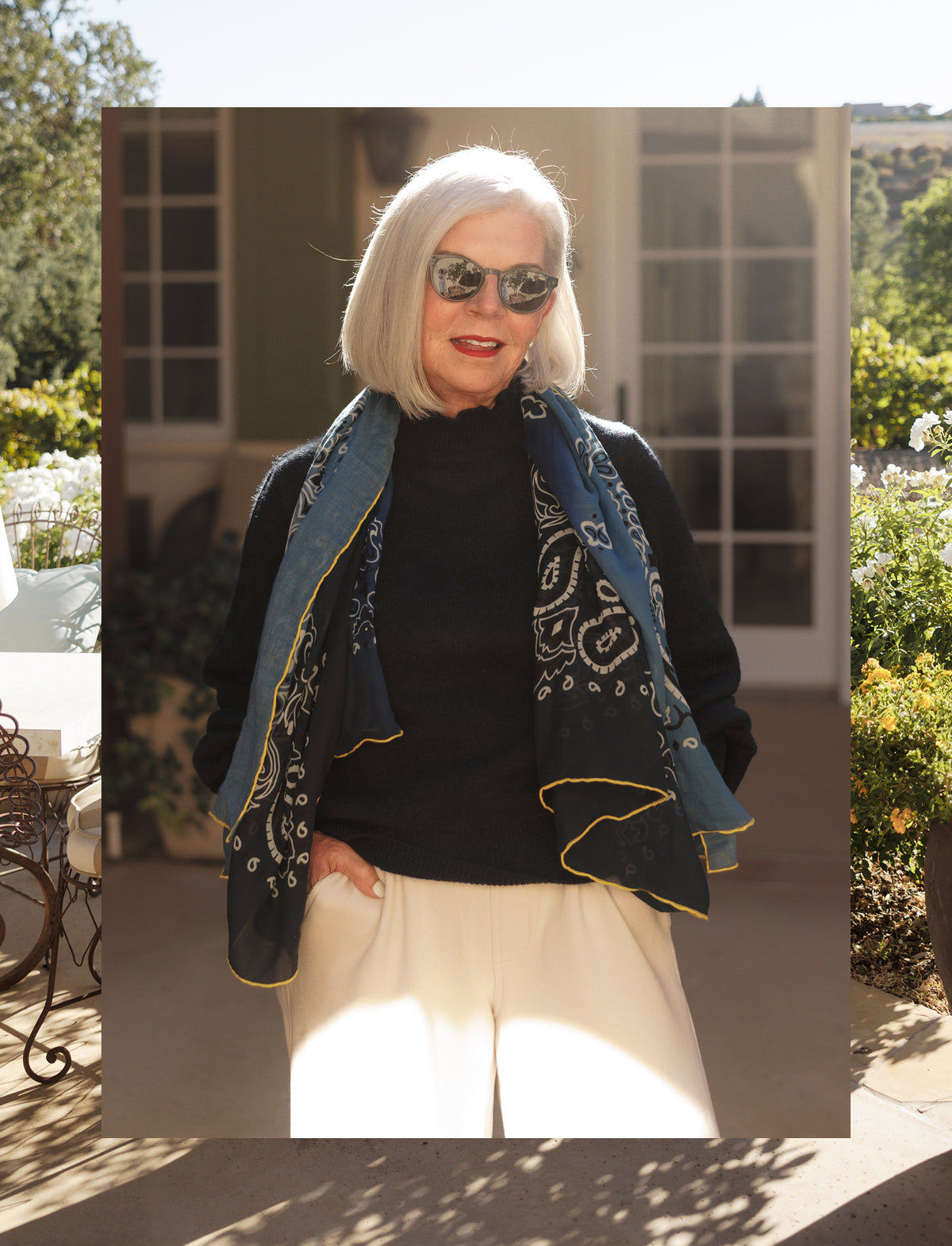 A woman with short white hair and sunglasses smiling while wearing a black knit sweater and a large navy blue and gold scarf draped over her shoulders. She is standing outdoors in what appears to be a garden setting.