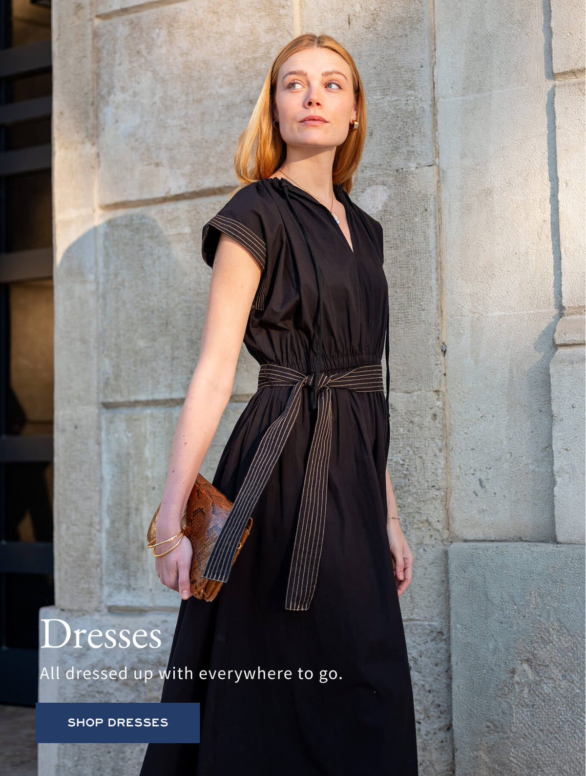 Promotional banner for women's dresses with a woman in a black dress and striped belt against a stone wall. Text: 'Dresses. All dressed up with everywhere to go. SHOP DRESSES'