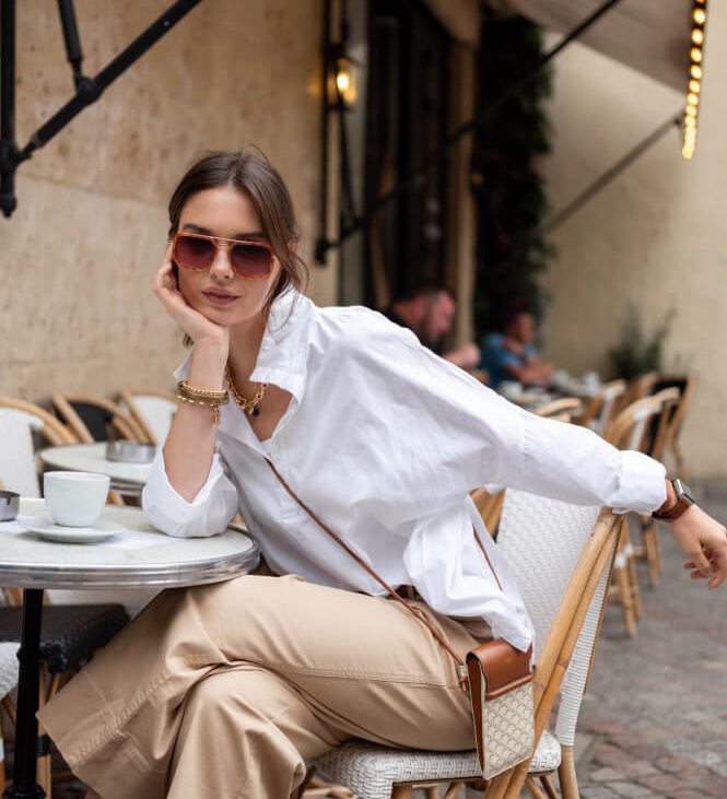 Image of a woman sitting at a cafe table wearing a white pop-up collar top with tan pants.