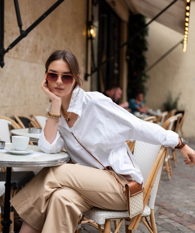 Image of a woman sitting at a cafe table wearing a white pop-up collar top with tan pants.