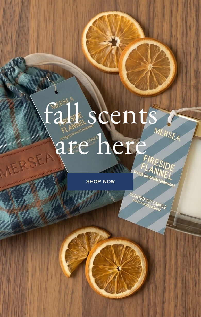 Promotional banner for fall scents. The image features a blue and green plaid bag and two product tags. Text: 'MERSEA fall scents are here. SHOP NOW. MERSEA FIRESIDE FIREWOOD [scent] [travel] [woodmoss]. SCENTRY CANDLE [scent] [luxury] [parfum].'