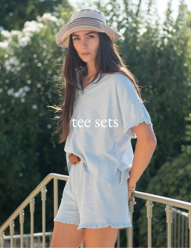 Woman wearing a light blue outfit with a hat, standing outdoors with greenery in the background.