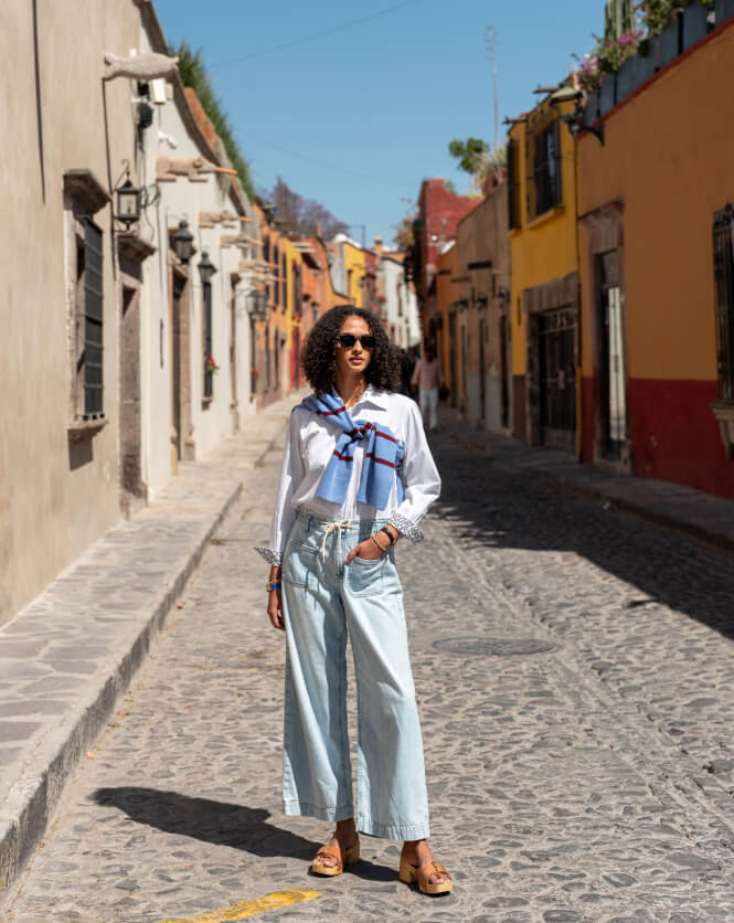 Image of a woman wearing a white button up shirt with a blue sweater draped over her shoulders and a light blue denim wide leg pants