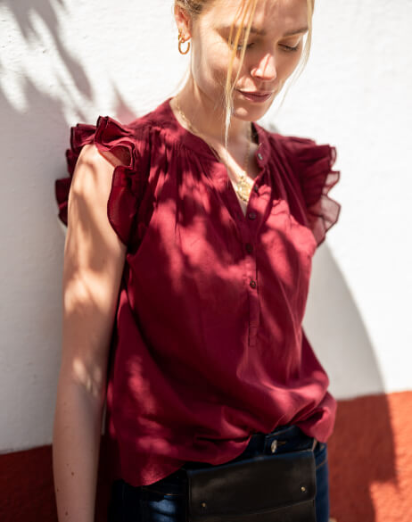 Woman wearing a burgundy blouse with ruffled sleeves against a white wall.