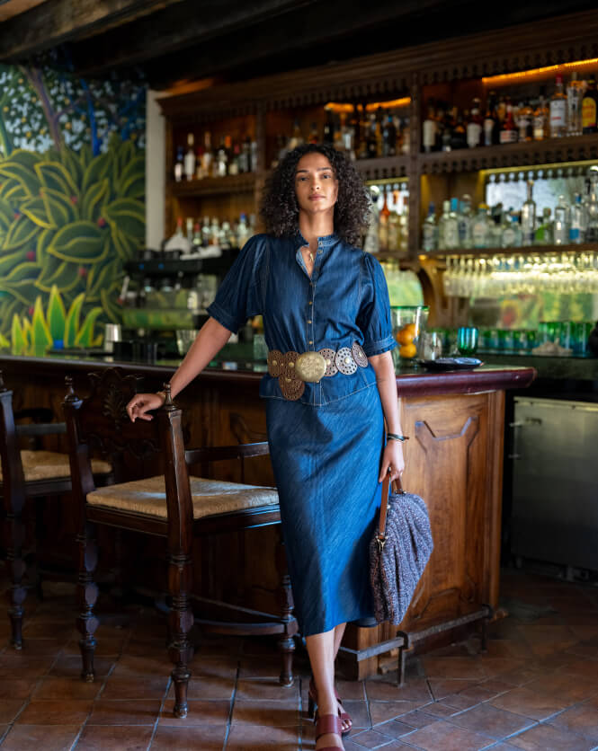 Woman in a blue dress standing in a bar with a wooden counter and shelves filled with bottles.