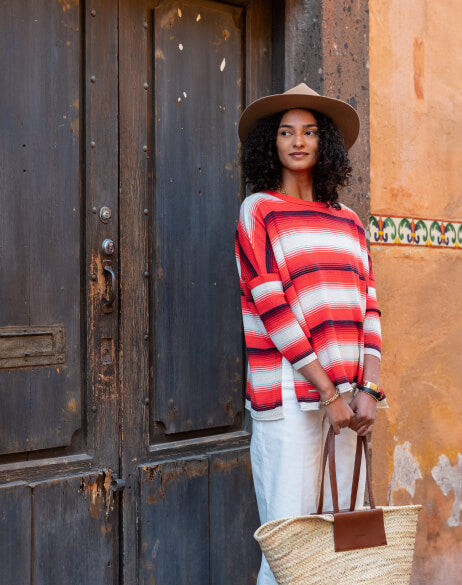 Woman wearing a red and white striped sweater and hat, holding a straw bag, standing in front of a rustic door.