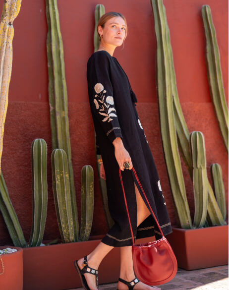 Woman in a black outfit with white patterns holding a red bag against a red wall with cacti.