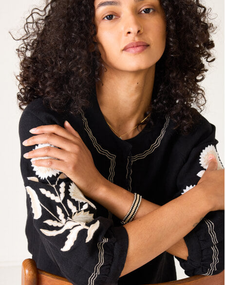 Woman wearing a black top with white floral patterns, sitting with arms crossed.