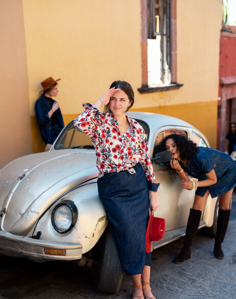 Woman in a floral blouse standing next to an old car with another woman inside, in a colorful street setting.