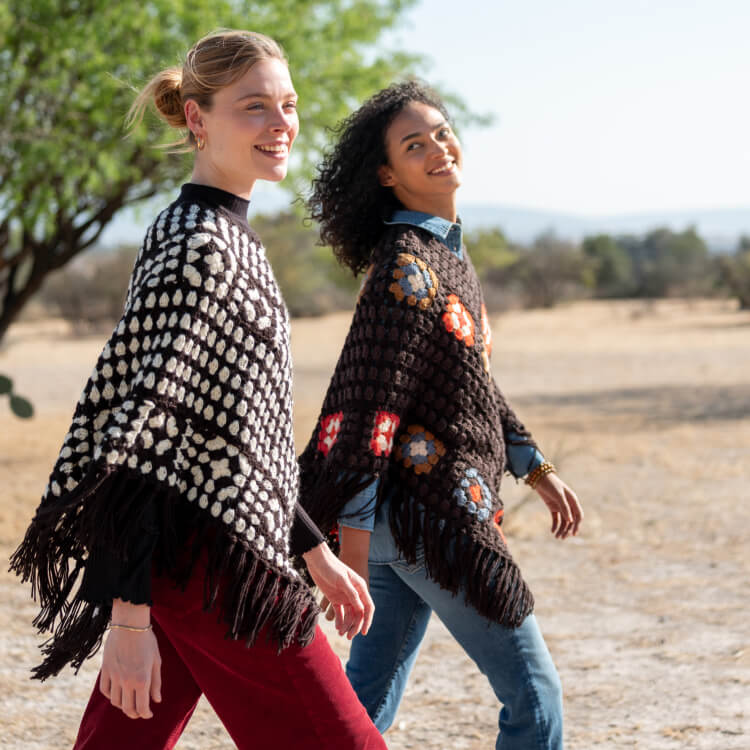 Two women wearing crocheted ponchos walking outdoors in a natural setting.