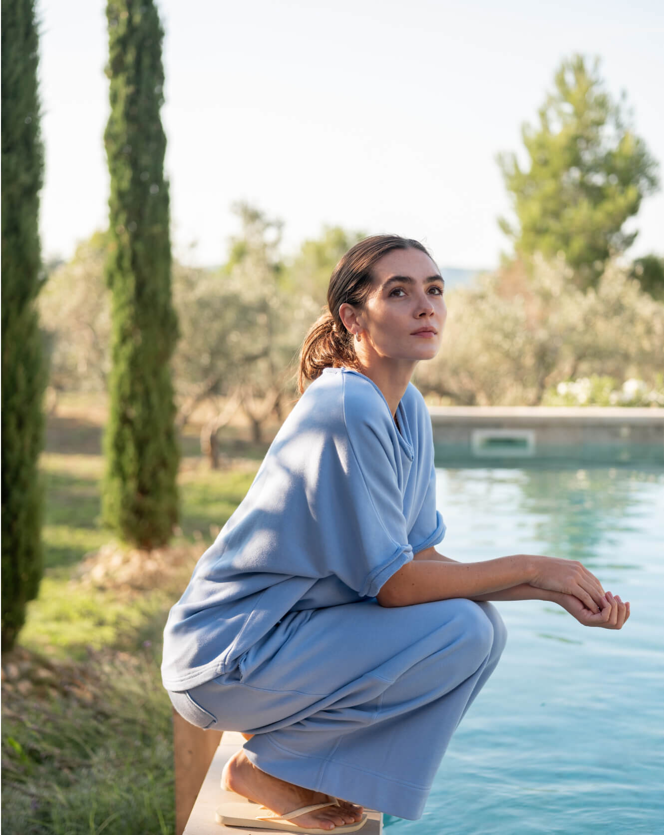 Woman in a light blue dress sitting by a pool with greenery in the background