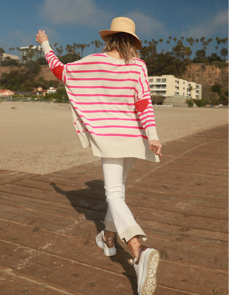 A woman in a wide-brimmed straw hat and a long pink and white striped tunic wearing white pants on a beach boardwalk.