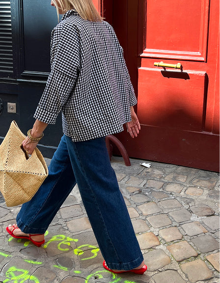 A woman in a black and white checkered oversized button-down shirt paired with wide-leg denim jeans and red loafers walks on a cobblestone street next to a red door while holding a woven basket.