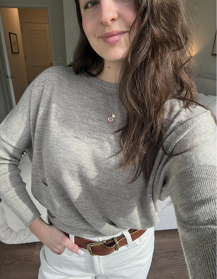 MERSEA ladies' travel-ready light gray ribbed knit sweater with a gold pendant necklace and a wide brown belt over white pants.