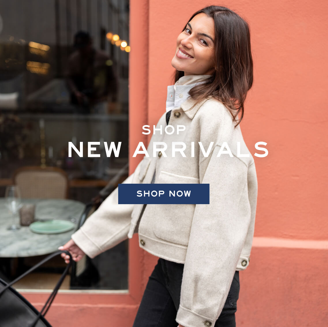 A woman in a cream knit cardigan smiling in front of a terracotta wall. Promotional banner. Text: 'SHOP NEW ARRIVALS SHOP NOW'