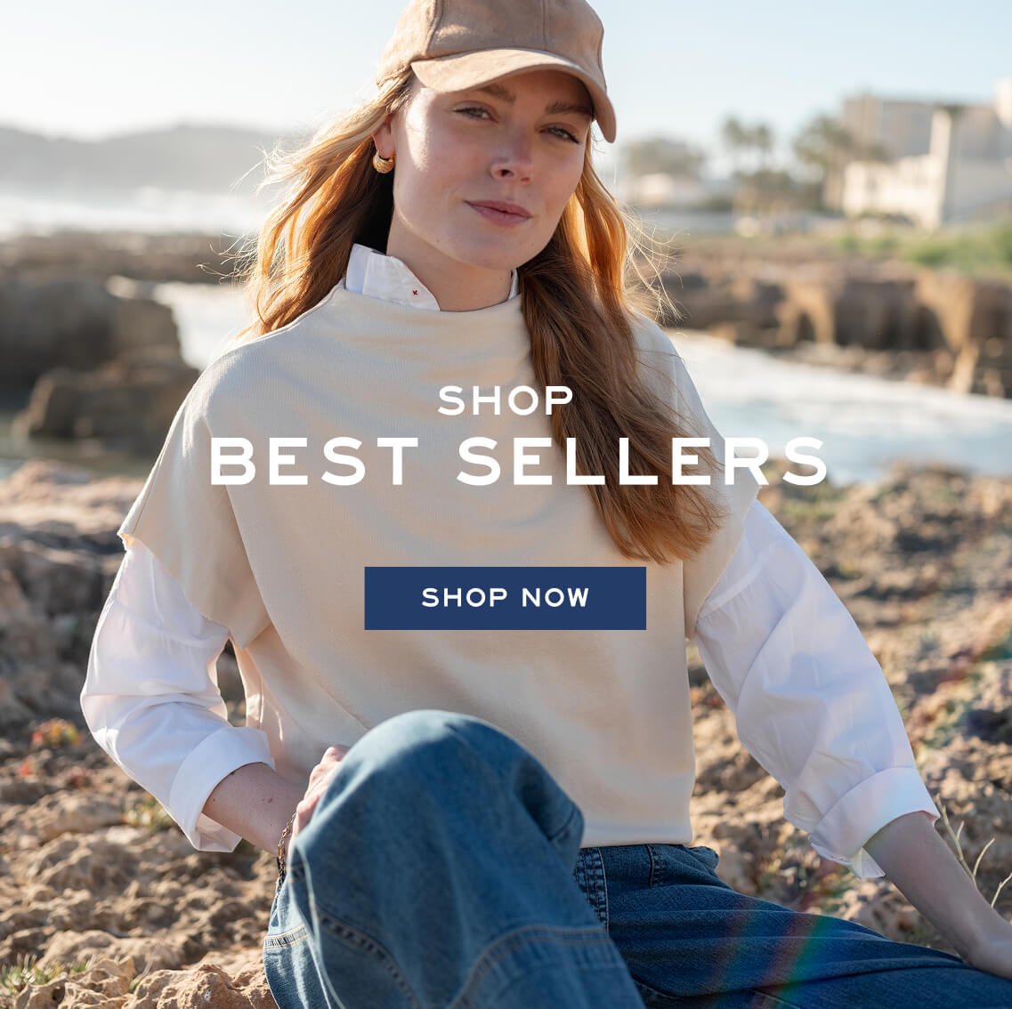 A woman in a beige cap and layered linen shirt sits on a rocky beach. Text: SHOP BEST SELLERS SHOP NOW