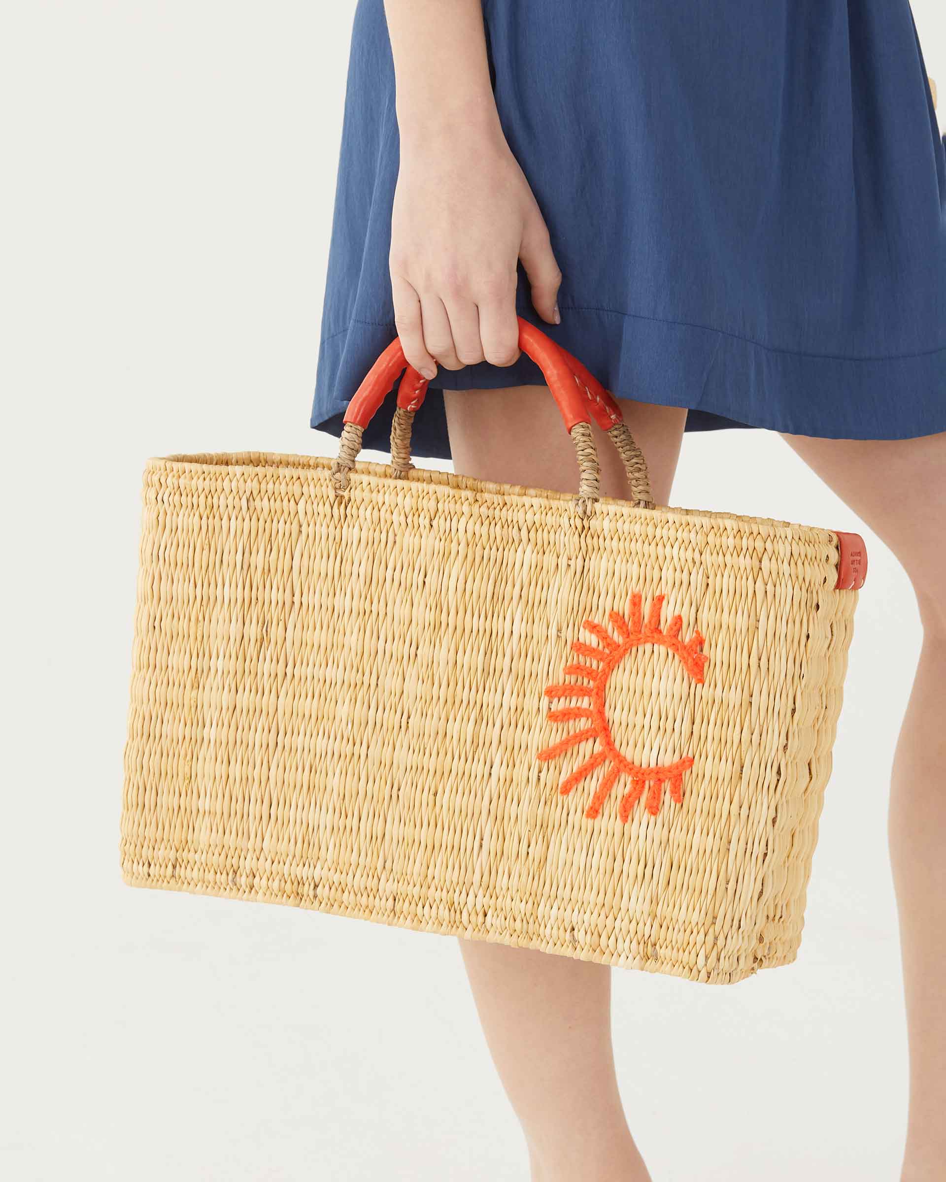 female holding straw bag with orange leather handles and a sun on a white background