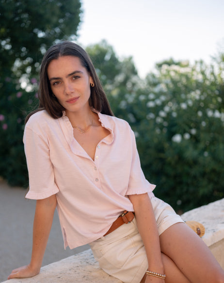 Woman in a light pink shirt and beige shorts sitting outdoors with greenery in the background