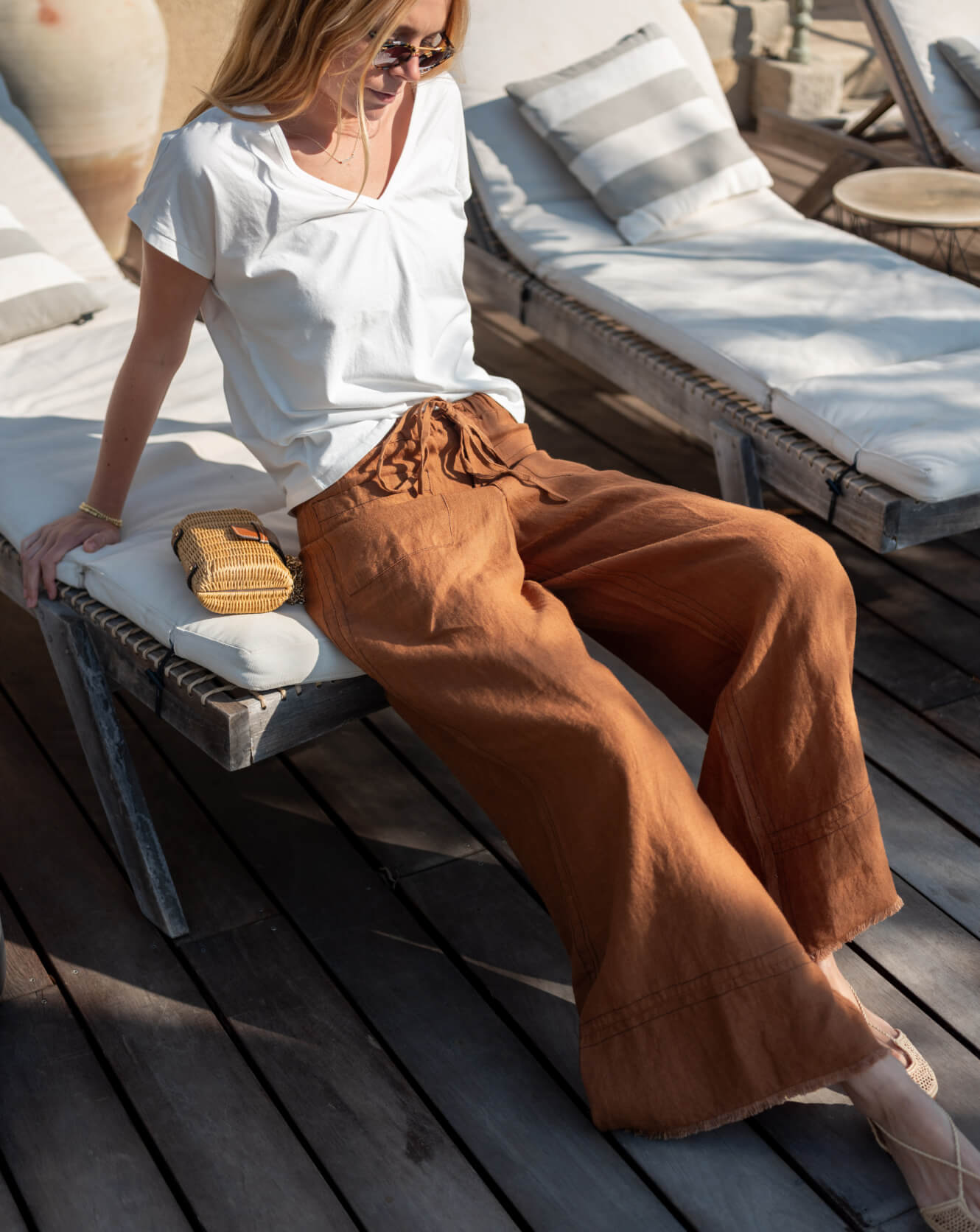 Woman sitting on a sun lounger wearing a white top and brown pants, with a wooden deck and outdoor furniture in the background.